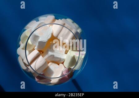 Mini marshmallows in un bicchiere di vino su sfondo blu. Marshmallows colorati in un calice di vetro. Vista dall'alto di un bicchiere pieno di dolcetti. Messa a fuoco selettiva. Foto Stock
