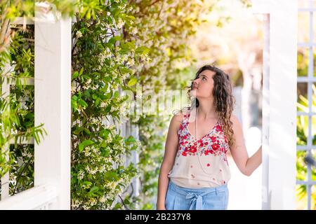 Ragazza giovane donna in piedi guardando in estate il giorno soleggiato in giardino toccando fiori di piante di vite fuori indossare pastello moda Foto Stock