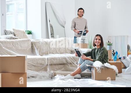 Bella giovane donna e suo marito con cornici che guardano a voi mentre disimballano le scatole nella loro nuova casa Foto Stock