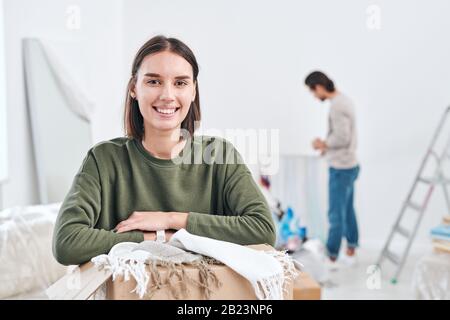 Felice giovane moglie con un sorriso toothy che ti guarda mentre disimballaggio le scatole sullo sfondo dell'uomo in piedi da parete Foto Stock