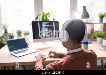 Vista posteriore del giovane IT-manager nelle cuffie premendo i tasti della tastiera mentre si trova davanti al computer portatile e al monitor del computer Foto Stock