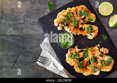 Bistecche di cavolfiore con salsa di lime coriandolo. Vista dall'alto su uno sfondo di pietra scura. Cibo sano, concetto di sostituto della carne a base di piante. Foto Stock