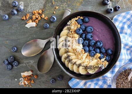 Ciotola di frullato di mirtillo sano con noce di cocco, banane, semi di chia e granola. Vista dall'alto della scena del tavolo su uno sfondo di pietra scura. Foto Stock