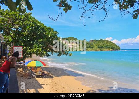 Castries, Santa Lucia - 23 novembre 2019. Bella giornata a Vigie turisti sotto ombrelloni multicolore spiaggia, signora locale che sta guardando loro Foto Stock