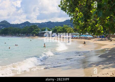 Castries, Santa Lucia - 23 novembre 2019. Turisti a Vigie Beach in una luminosa giornata estiva, alcuni sotto l'ombra di un FLUSSO sponsorizzato ombrelli Foto Stock