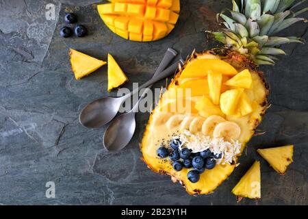 Ciotola sana di frullato in un ananas con cocco, banane, mango e mirtilli. Vista dall'alto su uno sfondo scuro di ardesia. Spazio di copia. Foto Stock