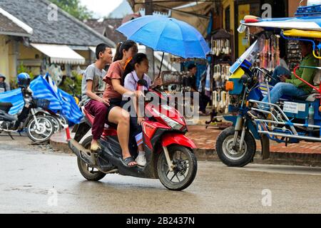 Tre persone con un ombrello in moto sotto la pioggia, Luang Prabang, Laos Foto Stock