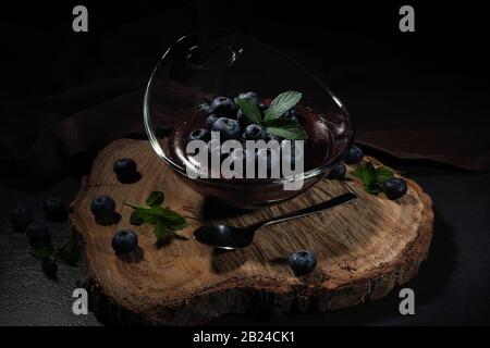 Budino al cioccolato con mirtilli, ben disposto in una ciotola di vetro su un piatto di legno Foto Stock