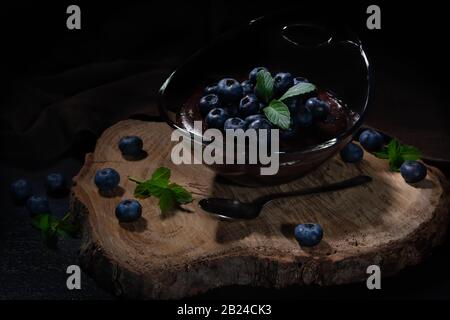 Budino al cioccolato con mirtilli, ben disposto in una ciotola di vetro su un piatto di legno Foto Stock