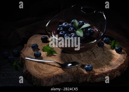 Budino al cioccolato con mirtilli, ben disposto in una ciotola di vetro su un piatto di legno Foto Stock