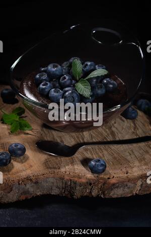 Budino al cioccolato con mirtilli, ben disposto in una ciotola di vetro su un piatto di legno Foto Stock