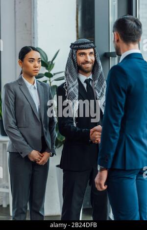 partner commerciali multietnici e sorridenti che si stringono a mano a una riunione con traduttori in ufficio Foto Stock