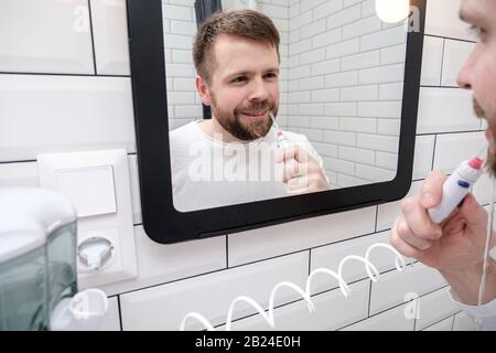 L'uomo contento pulisce i denti con l'aiuto di un irrigatore elettrico orale con un getto d'acqua, guardando nello specchio. Foto Stock