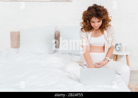 Interessante donna incinta che tocca il pancino e che usa un computer portatile a letto Foto Stock