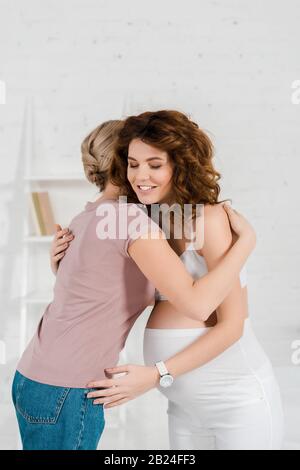 Donna incinta sorridente che abbraccia doula nel soggiorno Foto Stock