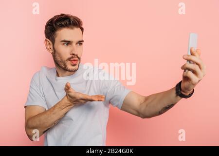 bel giovane uomo che soffia aria bacio mentre si ha video chat su smartphone su sfondo rosa Foto Stock