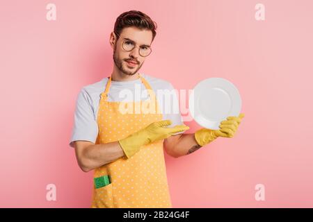 giovane uomo sicuro in grembiule e guanti in gomma che mostrano piastra pulita su sfondo rosa Foto Stock