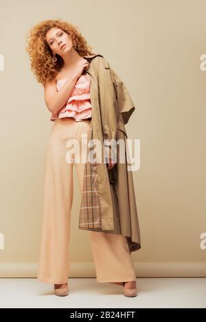 elegante donna redhead in top con volant e pantaloni che tengono trench mentre si posa su beige Foto Stock