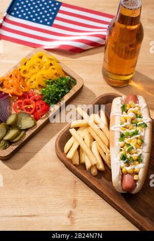 delizioso cane caldo vicino alla bandiera americana, tavola con verdure a fette, birra e patatine fritte su tavola di legno Foto Stock