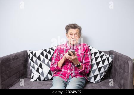 Tema vecchia persona utilizza la tecnologia. vecchio grigio-dai capelli donna caucasica con Rughe sorge casa in soggiorno sul divano e utilizza il telefono cellulare nelle mani, guarda Foto Stock