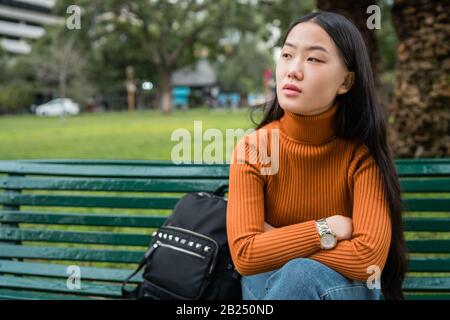 Ritratto di giovane bella donna asiatica pensierosa e seduta su una panchina nel parco all'aperto. Foto Stock