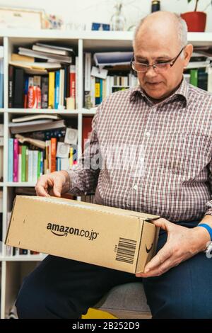 Parigi, Francia - 2 aprile 2019: Felice uomo anziano che detiene nuovo pacco è arrivato da Amazon Giappone con grande logotipo sul lato e mobili bibliotehque in background Foto Stock