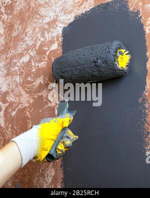 parete di casa dipinta a mano con vernice acrilica grigia con un rullo in guanti da lavoro gialli. Foto Stock