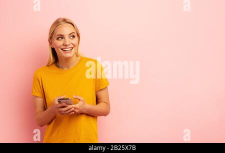 Bionda ragazza carina tipi un messaggio con il suo smartphone. Felice e sorpreso faccia di espressione. Sfondo rosa Foto Stock