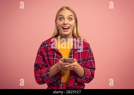 Bionda ragazza carina tipi un messaggio con il suo smartphone. Felice e sorpreso faccia di espressione. Sfondo rosa Foto Stock