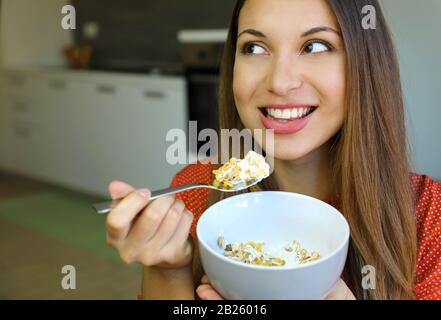 Primo piano di bella giovane donna che mangia skyr yogurt con cereali muesli frutta a casa, guardando al lato, concentrarsi sul modello occhi, immagine interna. H Foto Stock