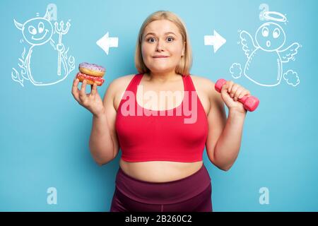 Fat ragazza pensa di mangiare ciambelle invece di fare palestra. Concetto di indecisione e dubbio con angelo e suggerimento del diavolo Foto Stock