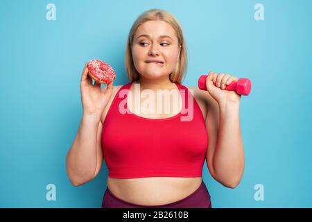 Fat ragazza pensa di mangiare ciambelle invece di fare palestra. Concetto di indecisione e dubbio Foto Stock