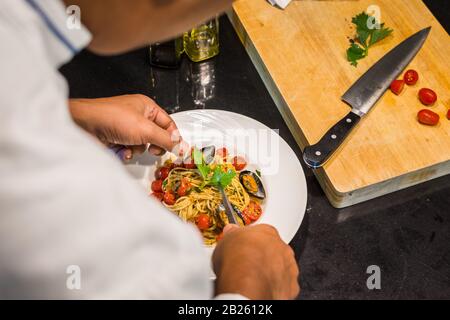Closeup di uno chef che serve e decorare un piatto spaghetti Vongole con pomodori roma e cozze accanto a una tagliabella e un coltello Foto Stock
