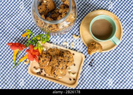 biscotti con pezzetti di cioccolato e noci decorati su un piatto di legno con una tazza di caffè, un vasetto di biscotti e fiori su un tovaglia sho bianco e blu controllato Foto Stock