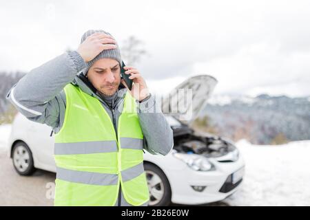 Uomo stressato in riflettente giubbotto chiamata servizio di assistenza stradale accanto a auto rotta con cofano aperto. Foto Stock