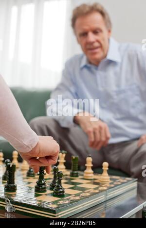 Uomo maturo che gioca a scacchi in un luminoso soggiorno - concentrarsi sulla mano della donna in primo piano Foto Stock