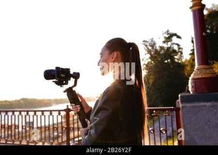 Attraente giovane brunetta che tiene lo stabilizzatore della macchina fotografica e che fa un film all'aperto al mattino. Sole che sorge sopra il fiume, estate caldo tempo e. Foto Stock