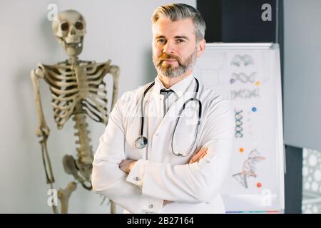 Dottore o professore di medicina con le braccia incrociate e stetoscopio intorno al collo guardando la fotocamera, in piedi in ufficio moderno. Scheletro umano sul Foto Stock