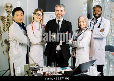 Gruppo misto-razza di giovani medici, operai di laboratorio, scienziati, con uomo maturo maschio supervisore, lavorando in laboratorio moderno, sorridendo e tenendo le braccia incrociate Foto Stock