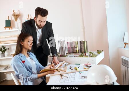 il team leader che prende appunti mentre si siede alla scrivania , la sua collega che ascolta il suo consiglio. coppia discutere i dettagli del lavoro di riparazione in ufficio wit Foto Stock