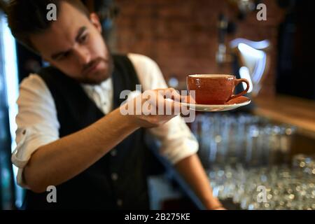 bel cameriere che guarda la tazza arancione e il soucer prima di dare loro un cliente. chiudi la foto. sfondo sfocato Foto Stock
