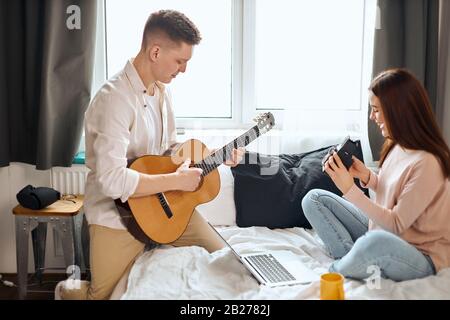 giovane famiglia di talento che si diverte in camera da letto, uomo che pratica canzoni di canto, coppia preparando il regalo creativo per i parenti in anniversario. creativ Foto Stock