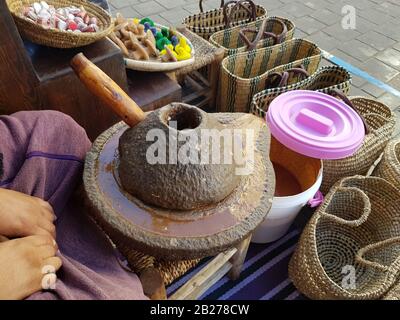 Al centro della composizione si trova una pressa per pietra per la produzione di olio argan. Si possono vedere le mani di una donna che produce olio di argan. Cestini di vimini, s Foto Stock