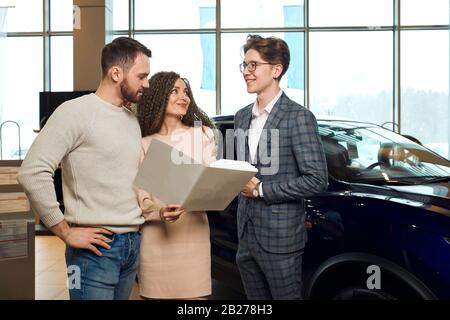 attraente coppia che acquista una nuova auto da un venditore, uomo che tiene cartella, documenti che parlano di caratteristiche del veicolo, vendita, sconto. primo piano foto Foto Stock
