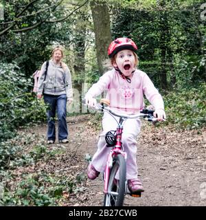 Una ragazza di sette anni fa si apre a tutta velocità attraverso i boschi su una bici rosa, sua madre camminando wearily dietro Foto Stock