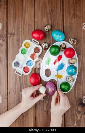 Uova pasquali dipinte a mano, pitture e pennelli su sfondo ligneo. Preparazione per la vacanza. Le mani delle ragazze disegnano un modello Foto Stock