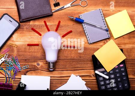 Cancelleria e lampada. Concetto di idea aziendale. Notebook, calcolatrice, clip colorate, biglietti da visita, portafoglio, penna e note adesive gialle su sfondo di legno. Lampadina a raggi rossi e utensili da ufficio. Foto Stock