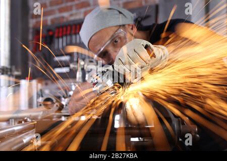 Uomo lavoro in officina di casa garage con smerigliatrice angolare, occhiali di protezione e guanti di costruzione, metallo levigatura fa scintille closeup, fai da te e del concetto di artigianato Foto Stock