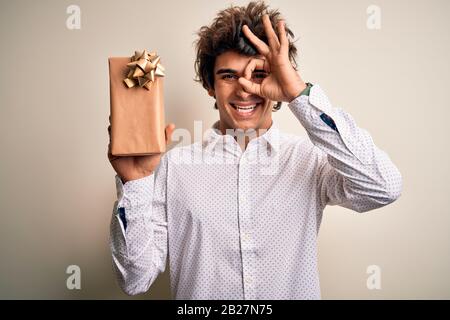 Giovane bell'uomo che tiene il regalo di compleanno in piedi sopra lo sfondo bianco isolato con il viso felice sorridente facendo segno ok con la mano sull'occhio che guarda thug Foto Stock