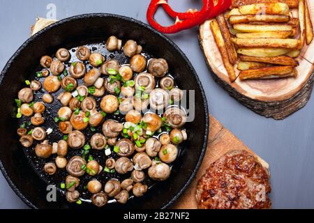 funghi d'oro champignon cosparsi di cipolle verdi in una padella nera in ghisa patatine fritte e succosa cotoletta di carne sulla foresta di cerotti in legno su un dorso grigio Foto Stock